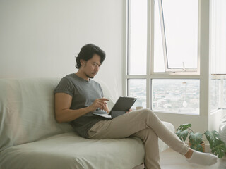 Fototapeta premium Asian man working on his laptop sitting on the sofa in the living room, feeling calm and relax with his work at home.