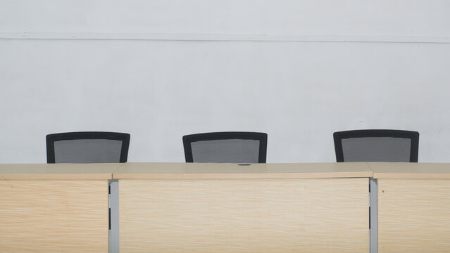 A row of empty black ergonomic mesh chairs lines a long wooden table in a clean, modern conference room, ready for a meeting