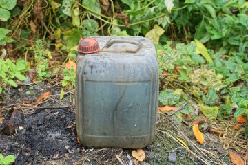 One old gray dirty plastic canister with a red cap filled with black liquid stands on the ground on a summer street