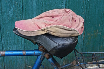 one red black cap made of fabric lies on the black leather saddle of an old bicycle near a green...