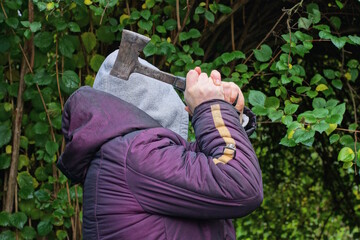 An aggressive man in a purple jacket and a gray hoodie swings an axe in his hand over his head against the backdrop of green vegetation on a spring street