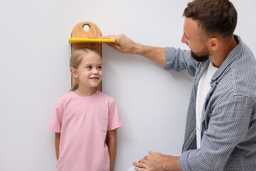 Father measuring height of his little daughter with scale near light wall indoors