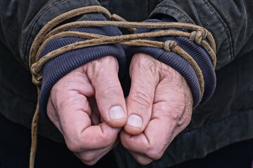 The hands of a man in black and blue clothes are tied with a brown rope on an autumn street