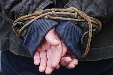The hands of a man in black and blue clothes are tied with a brown rope on an autumn street