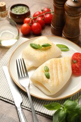 Pieces of tasty grilled squid, tomatoes, basil and cutlery on wooden table, closeup
