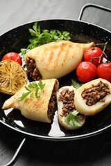 Pieces of tasty grilled squid stuffed with mushrooms, parsley, tomatoes and lemon in pan on dark textured table, closeup