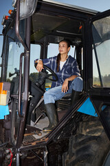 Naklejka premium Young adult Caucasian woman sitting in tractor cab holding steering wheel looking forward outdoors on farm field showing confidence and agricultural work involvement
