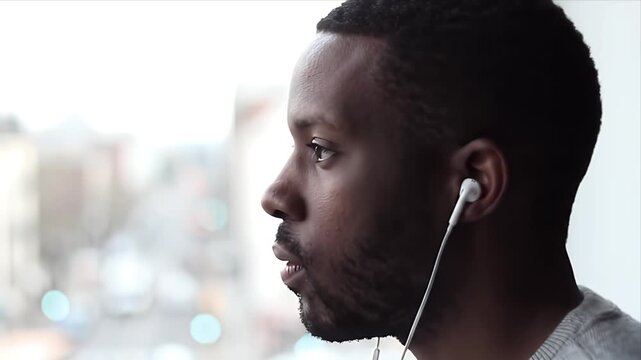 Relaxed Young Black Man Thoughtfully Listening to Music with Earphones by a Bright Window Pensive Millennial Gazing at Blurred Urban City View