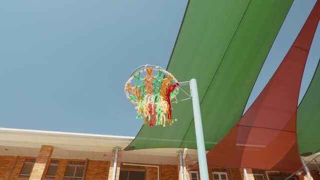 A basket ball being thrown and missing the colourful basketball net with the blue sky and green and red shade sails in the background.