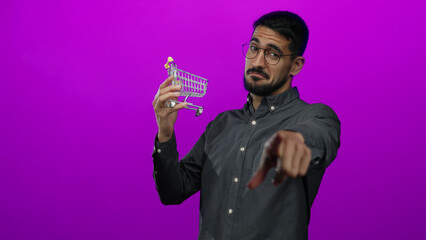 Young man with glasses holding mini shopping cart against pink background, smiling and pointing, creating a humorous retail concept with confident expression.