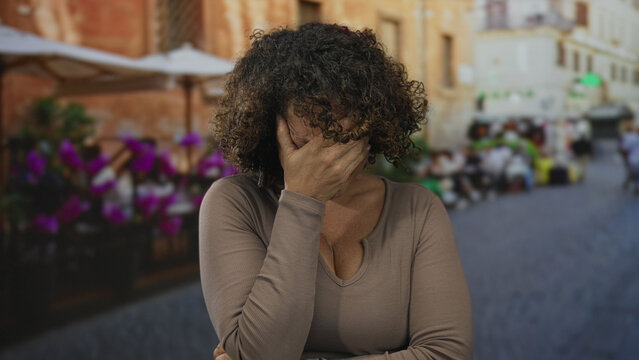 Hispanic woman covering face with hand at outdoor restaurant terrace beside flower pots and tables; sadness.