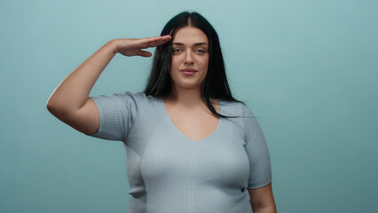 Woman saluting against an isolated blue background showcasing confidence and style with her plus...