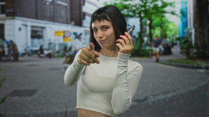 Fototapeta premium Woman wearing white croptop holds vaporizer and smiles in cobblestone street; carefree confidence.