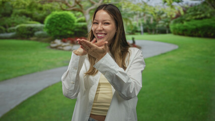 Fototapeta premium Woman smiling and blowing a kiss with cupped hands on a park path surrounded by green lawn and trees; joy playfulness warmth.