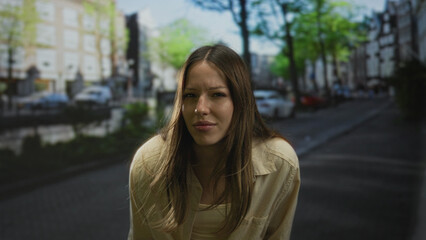 Fototapeta premium Woman squinting and leaning forward with windblown hair and beige shirt on city street, face centered and engaging the camera; curiosity.