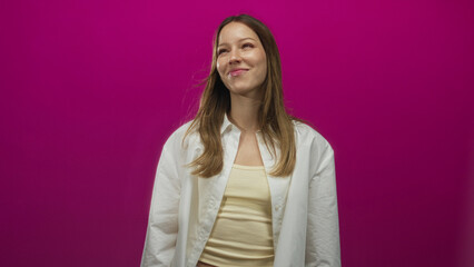 Fototapeta premium Woman in white shirt and cream tank top smiles and looks up in magenta studio; playful warm curiosity.
