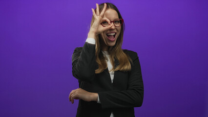 Woman making ok sign with right hand over eye wearing red glasses and black blazer in purple studio; playful confidence.