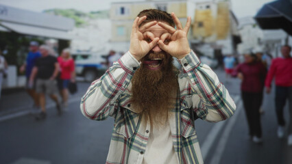 Young man with long beard makes binocular gesture with hands over eyes on sunlit busy street; curiosity.
