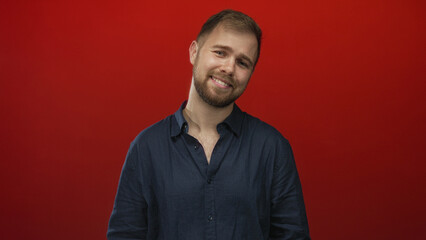 Obraz premium Man smiling with visible face and tilted head, beard and open collar, posed in red studio; cheerful optimism.