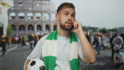 Man holding soccer ball and phone to ear in front of coliseum building, wearing green scarf and white shirt, looking upward; team loyalty anticipation.