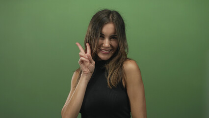 Fototapeta premium Woman shows peace sign hand and bright smile towards camera in green studio setting, hair framing face and casual posture; joy.