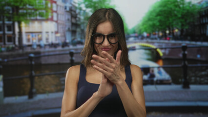 Fototapeta premium Woman smiling and clapping hands with bare arms wearing glasses on amsterdam street by canal; celebratory joy.