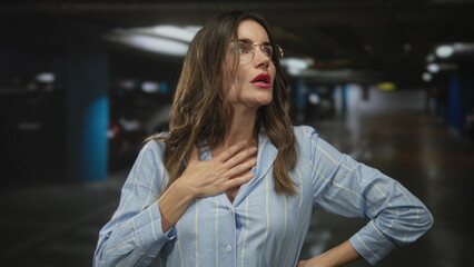 Fototapeta premium Woman wearing glasses holds neck next to car in an underground parking garage looking concerned; anxiety.