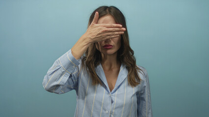 Woman covering her eyes with her hand against a blue studio backdrop while wearing a striped shirt; mystery.