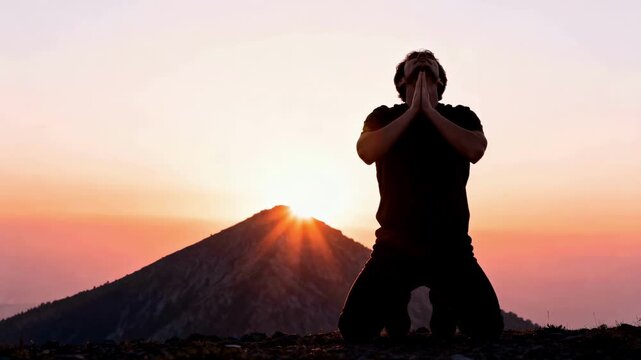 Silhouette of a man kneeling in prayer on a mountain at sunrise. A person worshiping with raised hands against a dramatic sky. Faith and spirituality concept