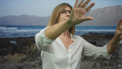 Woman hand reaching upward through sea breeze with waves rolling behind on street wearing white shirt and glasses; serenity.