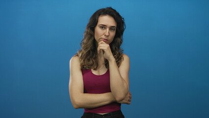 Fototapeta premium Young hispanic woman standing with arms crossed and hand on chin against blue studio wall; contemplation.