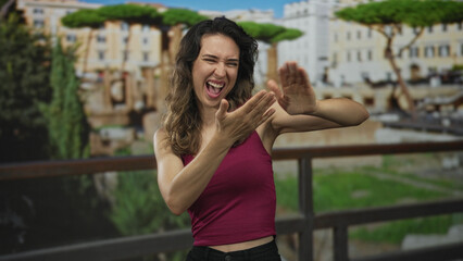 Woman flicking hand gesture at roman ruins building under bright sunlight in outdoor setting; exuberance.