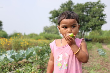 A heartwarming portrait of a young Indian girl standing in a rural farm setting, eating a slice of fresh green fruit. Dressed in a pink top, she looks away thoughtfully against a soft-focus background