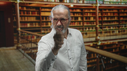 Man points finger at camera in library building among bookshelves with clenched jaw and raised hand  anger confrontation authority. © Krakenimages.com