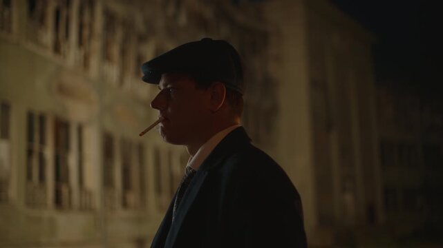 Side view of a confident man in a retro flat cap and coat smoking a cigarette on a dark city street, with smoke rising in the night air against a blurred architectural background from the 1920s