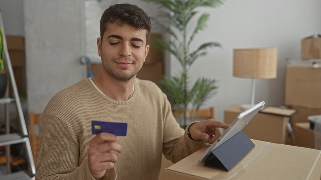 Man holds card after pointing around home as hispanic guy finishes moving purchase checkout unpack tasks.