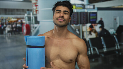 Man stands shirtless holding rolled yoga mat against bare chest near checkin counter in airport terminal; fitness travel anticipation.