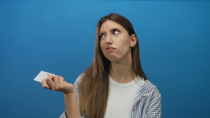 Young hispanic woman holding white plastic card with finger on chin against blue background in studio; uncertainty.