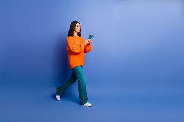 Naklejka premium Young woman in orange sweater using smartphone while walking on blue background showcasing modern and casual style