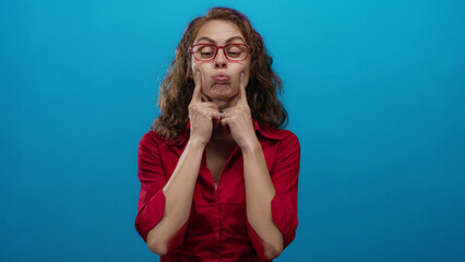 Woman with curly hair and red glasses wearing a red shirt making playful faces against a vibrant blue background, expressing joy and humor.