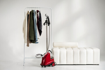 A light autumn capsule wardrobe on a rail hanger in a white room with a modern ottoman. A clothes steamer stands next to the hanger. Mockup with copy space.