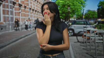 Fototapeta premium Young hispanic woman wearing black tee covers mouth with hand on sunlit street while smiling broadly; carefree joy.