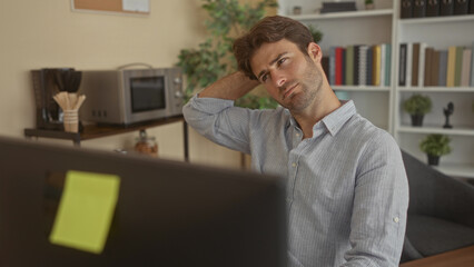Man with hand behind neck at computer monitor in office workplace, seated at desk and leaning back...