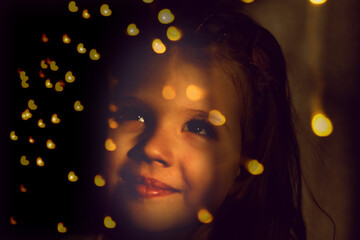 Happy smiling girl looking into the window decorated string lights. The Christmas flares lights...