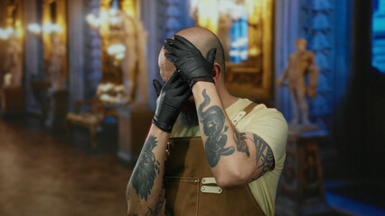 Bald man with gloves and tattoos stands indoors in a museum setting, conveying a moment of stress or contemplation amidst classical decor.