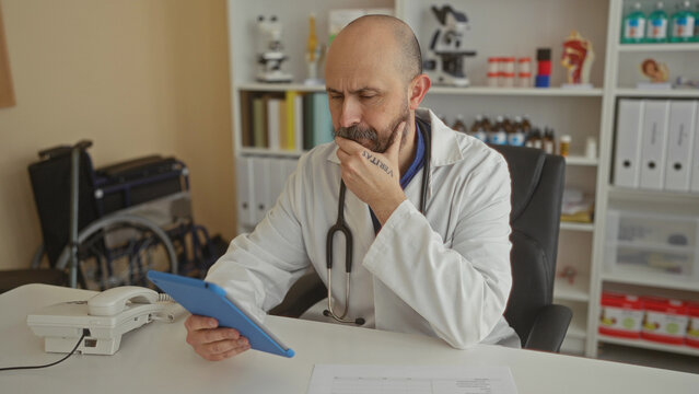 Bald man with a beard in a clinic examining a tablet, surrounded by medical equipment inside a hospital room. - Powered by Adobe