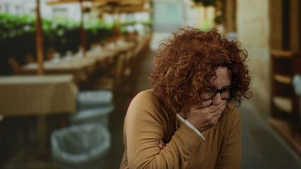 Woman laughing on an outdoor terrace in a restaurant, showcasing humor with tables and greenery in the background, wearing glasses and a brown sweater reflecting candid joy.