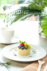 A delectable fruit tart on a decorative white plate, featuring fresh berries and kiwi, next to a cup of beverage
