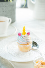 A whimsical unicorn cupcake on a decorative plate, set on a bright table with a spoon and a cup of tea. It's a delightful treat perfect for any occasion.