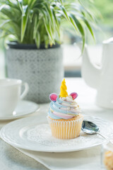 A cute unicorn cupcake on a plate next to a cup, teapot and plant. The cupcake is in the shape of a unicorn and is decorated with icing and a horn